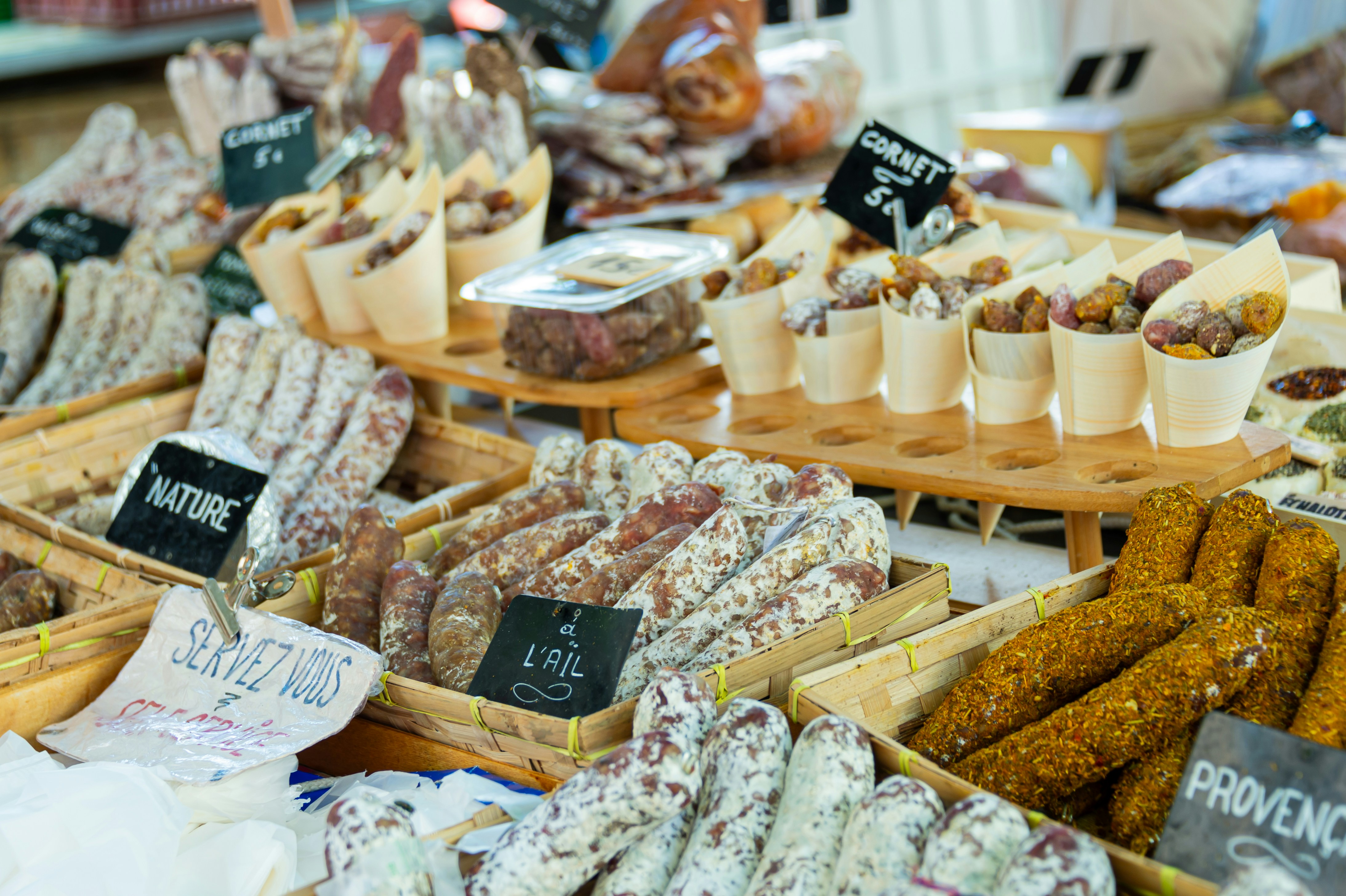 A French market counter stacked with saucisson in baskets and hand-chalked signs reading Nature, L&rsquo;Ail and Servez Vous