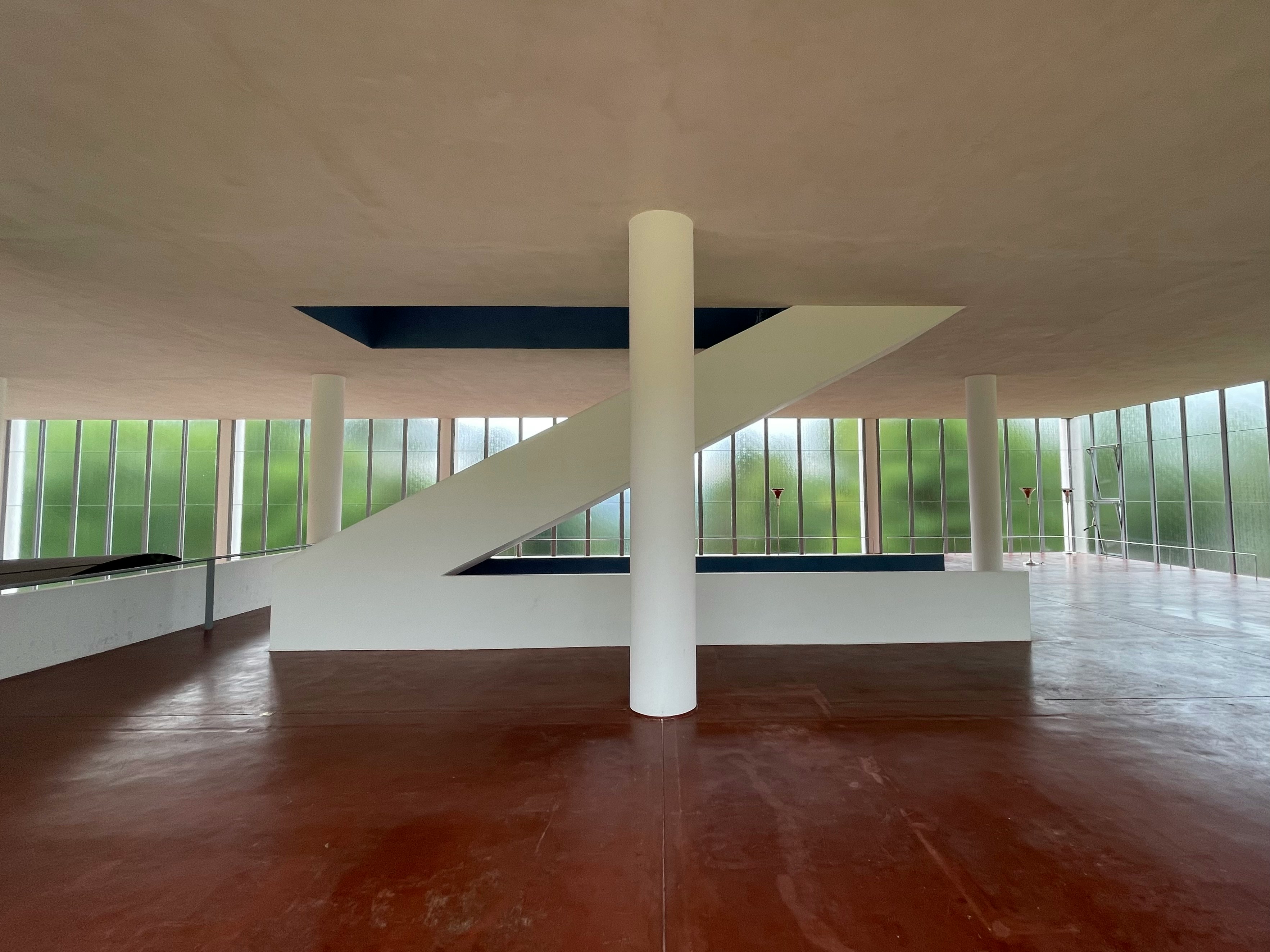 An empty modernist interior: a white diagonal stair against a ribbon window, oxblood floor, single column